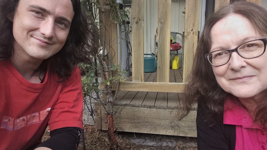 A man and a woman crouched in front of a narrow hole under their house, both smiling to camera.