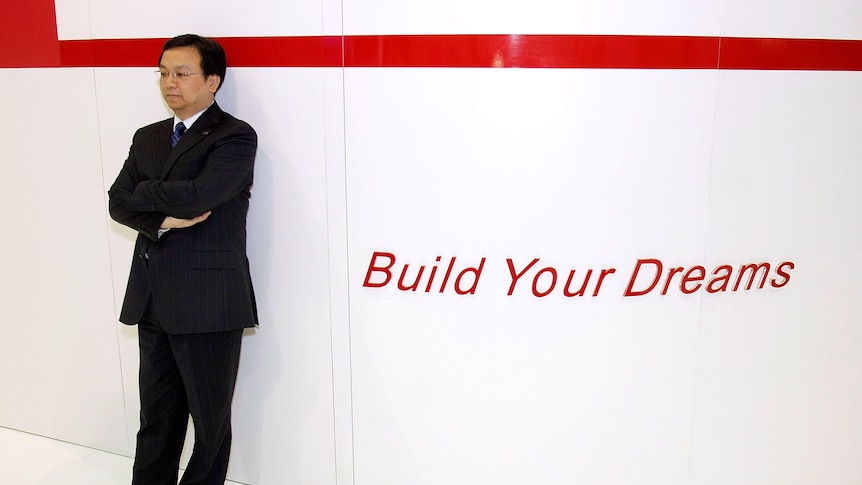 A middle-aged Chinese man in a black suit standing next to a sign that says 'Build Your Dreams'