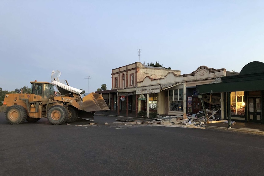 A front-end loader stands in front of a shop that is badly damaged.