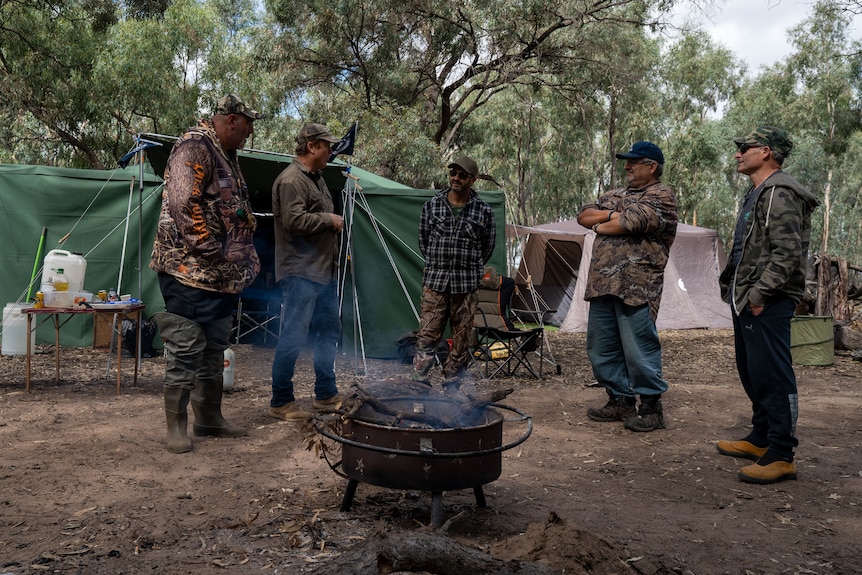 A group of men wearing camouflage clothing standing around a fire with tents in background