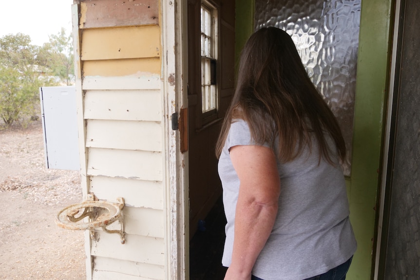 a woman with long hair, gray t-shirt and jeans looks at her newly purchased wooden house that cannot be lived in
