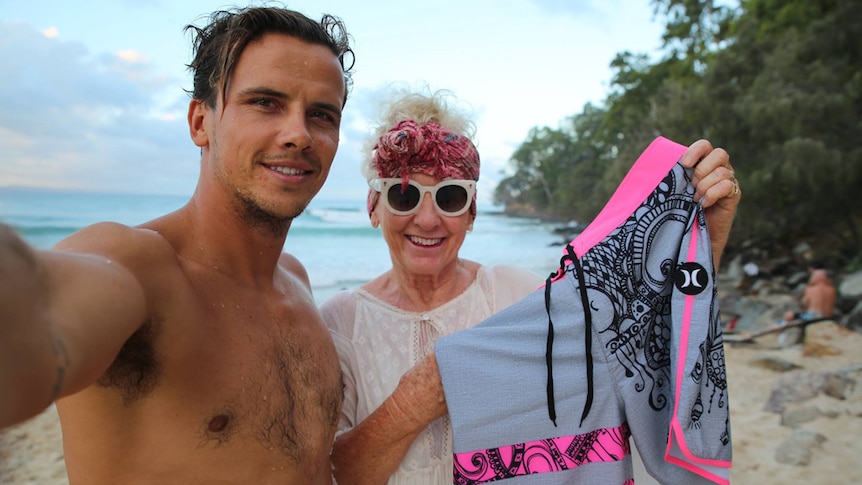 Son and mother at the beach holding up pink board shorts, smiling.