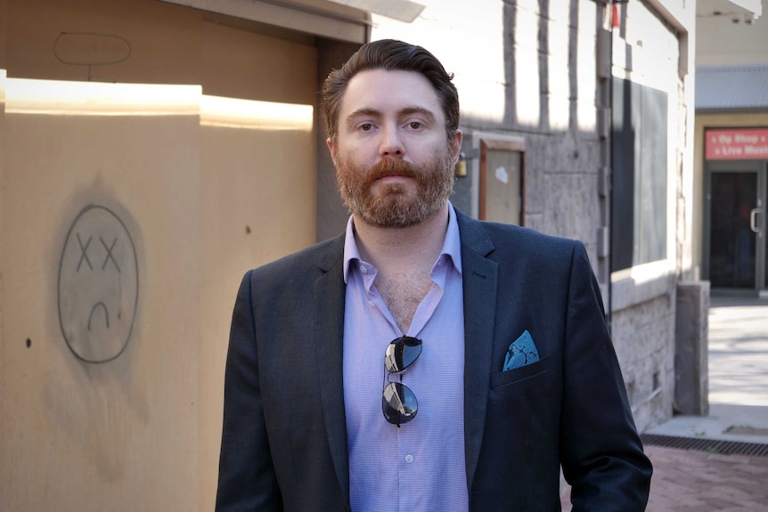 A man in a suit and casual business shirt stands in an alleyway with a boarded up shop in the background