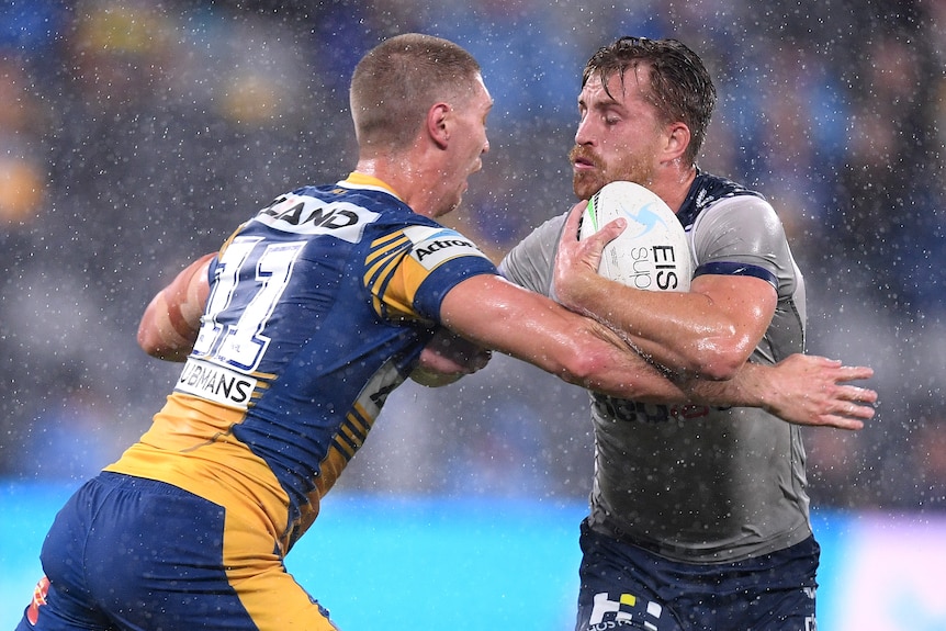 A Melbourne Storm NRL player holds the ball with his left arm as he palms off a Parramatta opponent.