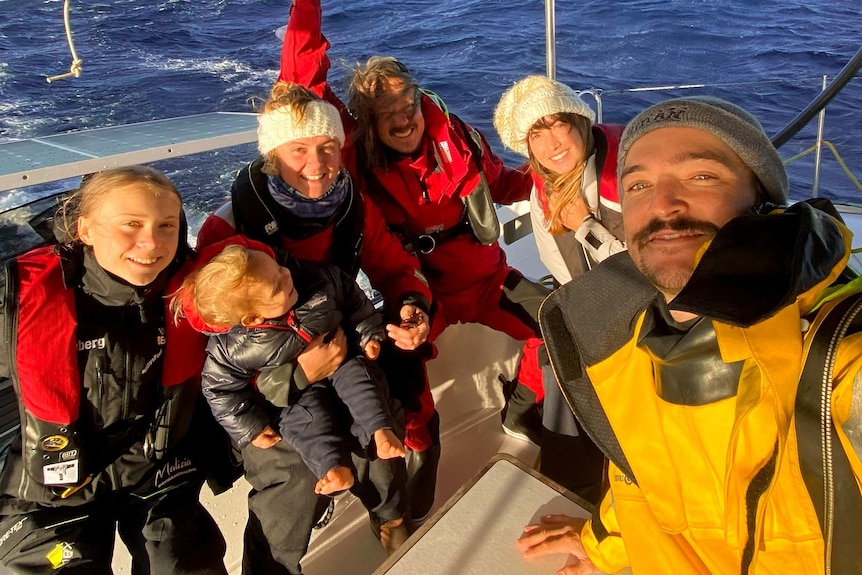 Greta Thunberg and a group of people pose for a photo smiling and wearing life jackets, on the deck of a catamaran at sea.