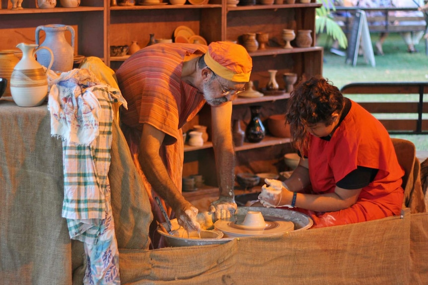 A man dressed in robes shows a young girl how to make a cup on a pottery wheel.