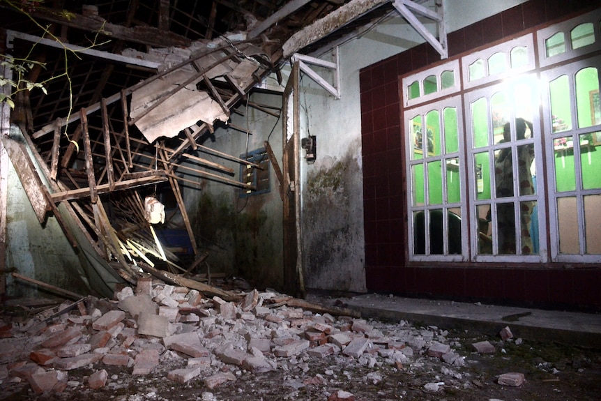 A woman surveys a part of her house damaged by an earthquake as debris lays on the floor and parts of the ceiling have fallen.