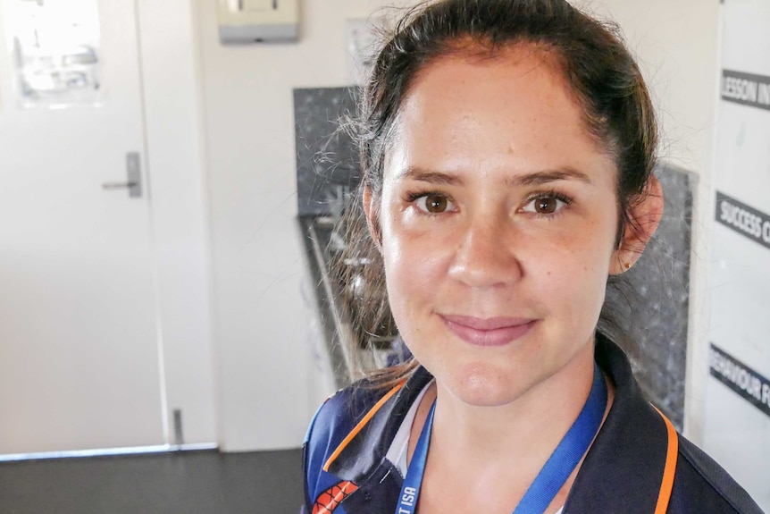 A young woman with brown eyes and long brown hair, wearing a black and orange polo shirt, smiles.