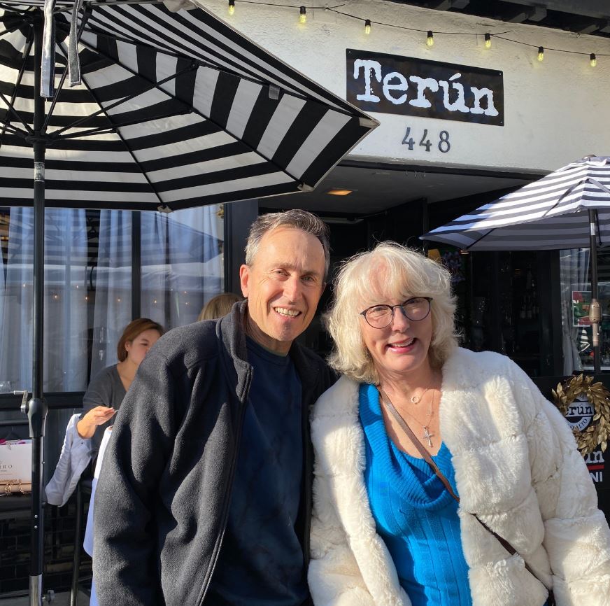 A man wearing a jumper and a woman wearing a white fur coat smile together in front of a cafe.