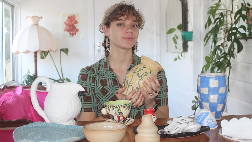 A young woman with curly hair holds a ceramic vase in her hands. Other handmade pottery pieces are on the table in front of her.