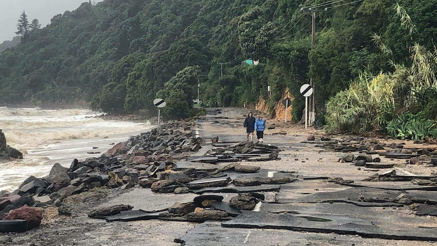 Rocks and debris litter a road near Te Puru, after the storm made waves to crash over it. Two people walk down the closed road.
