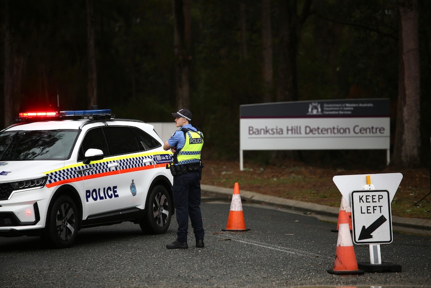 A police officer stands next to a police car at the entry to Banksia Hill Juvenile Detention Centre, with a sign on the verge.