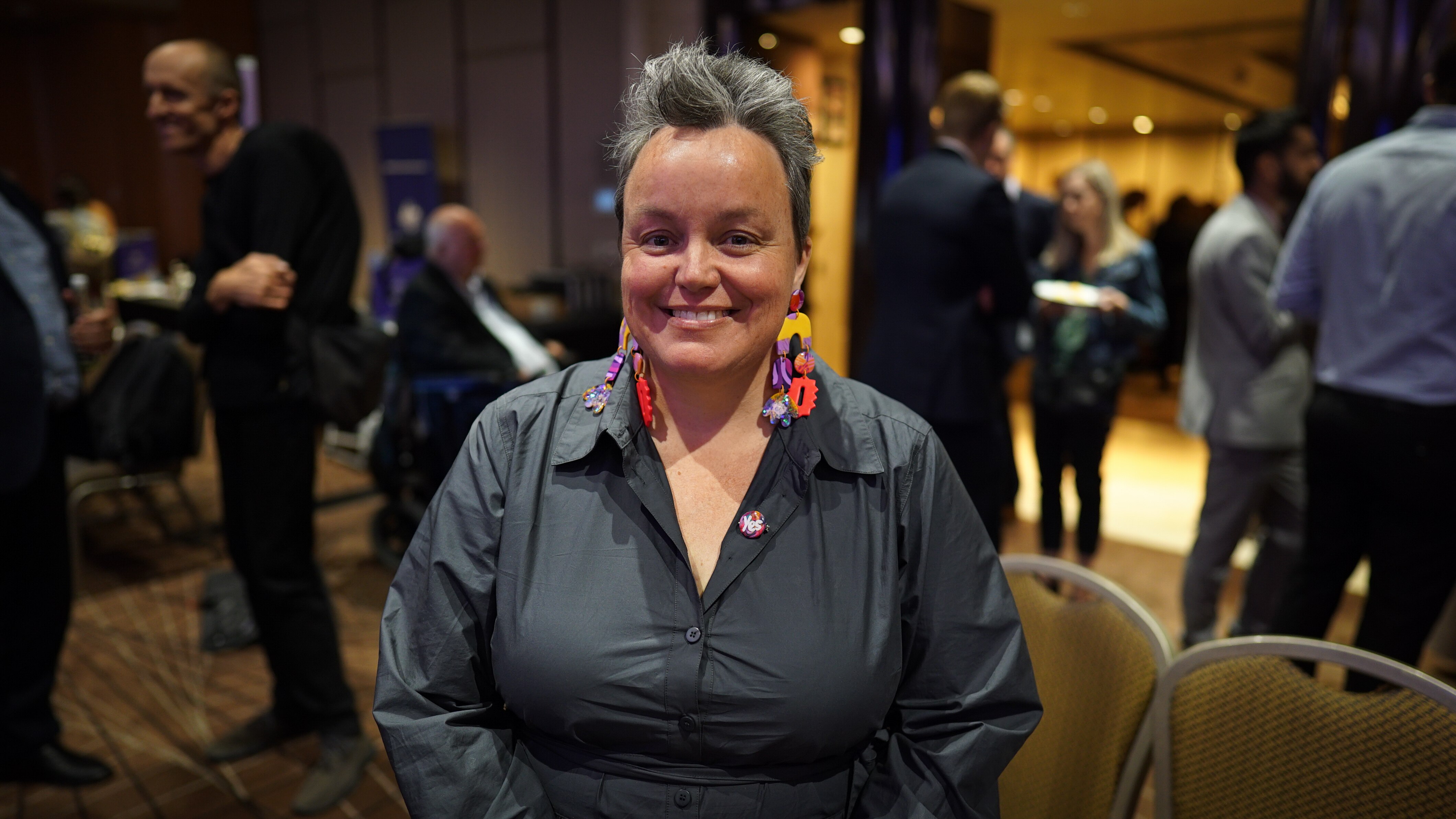 A woman with short grey hair and long earrings. She is smiling and standing in a ballroom