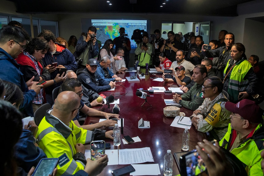 A group of people sit around a table, holding phones while wearing hi-vis