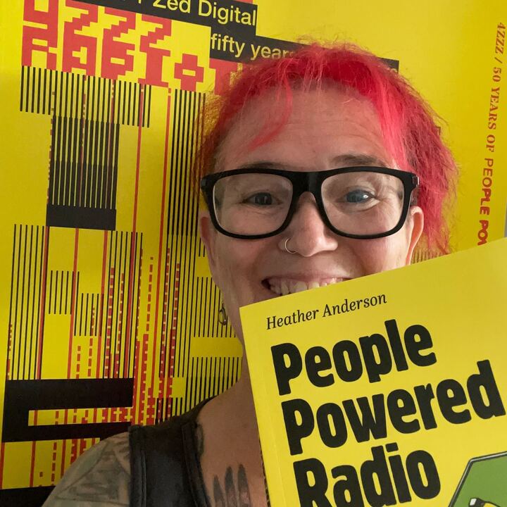 A smiling older woman with red hair & glasses takes a photo with her yellow-jacketed book cover