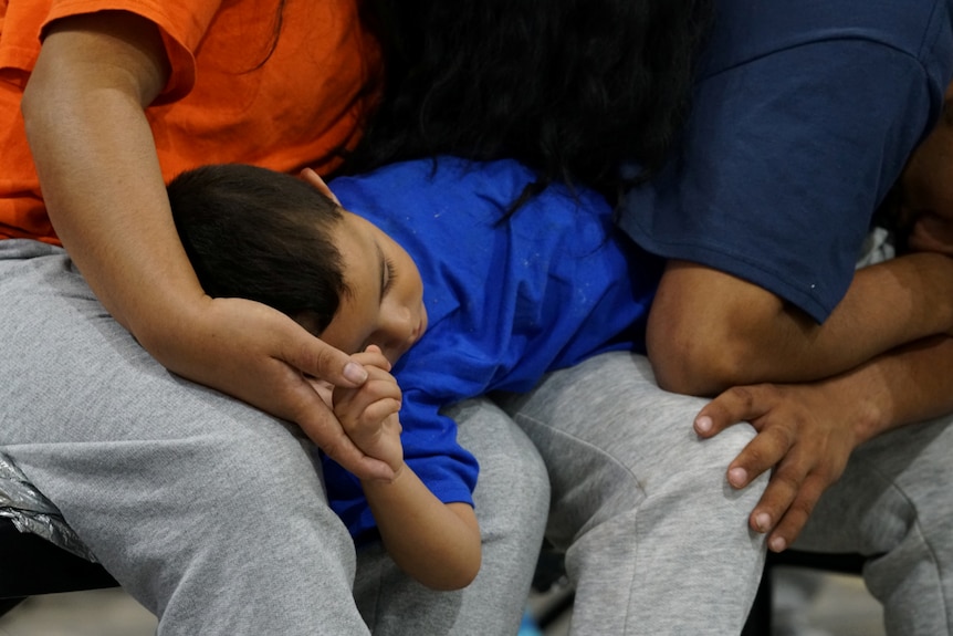 A sleeping boy holds his mother's hand while curled up in her lap