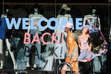 A young man wearing a mask walks past a store with the words "welcome back" painted on its door.