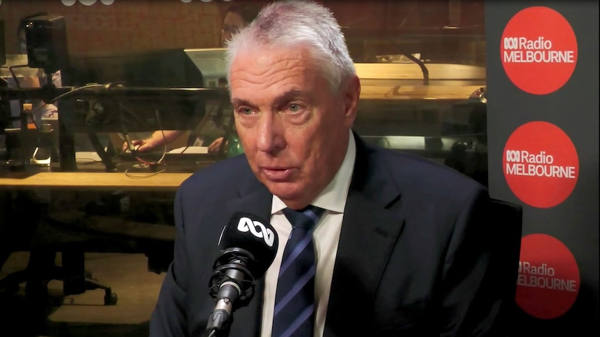 A man in a blue suit and tie sits in a radio studio in front of the microphone. 