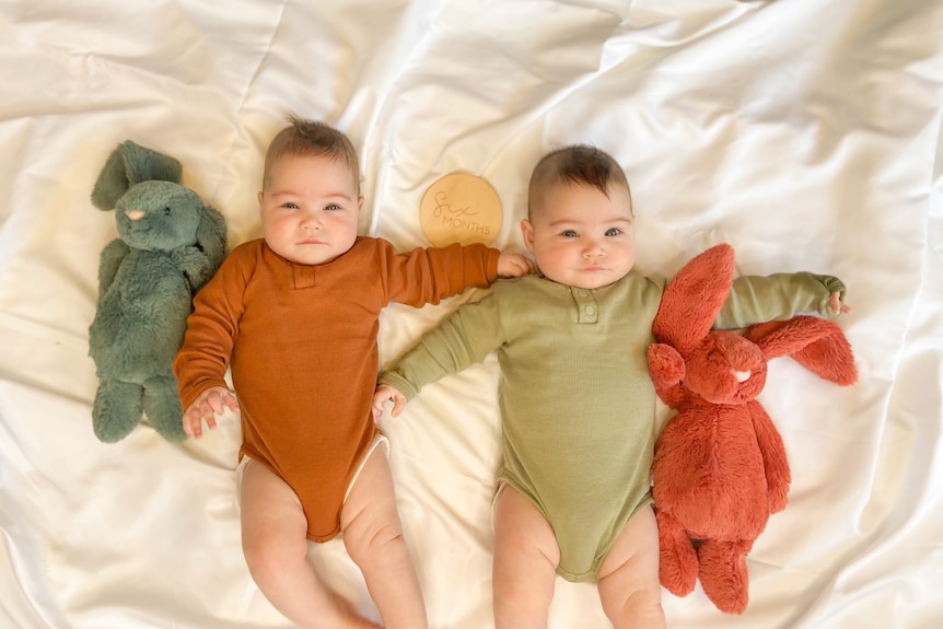 Twin babies Mia and Ava lying on a white sheet with two big toy bunnies, and a little marker saying "six months".