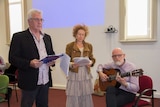 A man and woman standing holding papers or folder and another man sitting playing a guitar