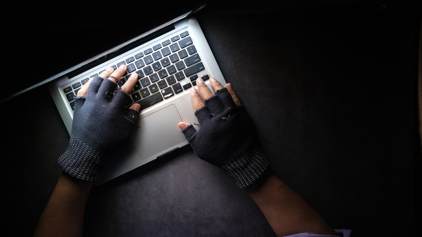 A pair of hands wearing fingerless gloves using a laptop computer in a dark room.