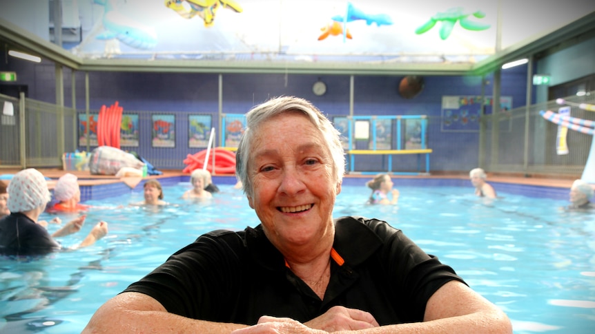 An older woman in a swim t-shirt leans her folded arms on the edge of a pool, in the background others frolic behind her.