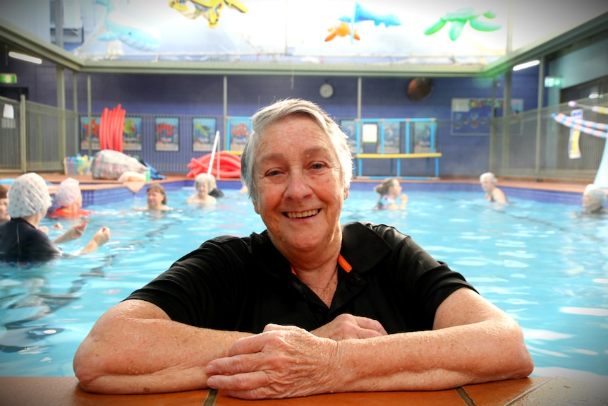 An older woman in a swim t-shirt leans her folded arms on the edge of a pool, in the background others frolic behind her.