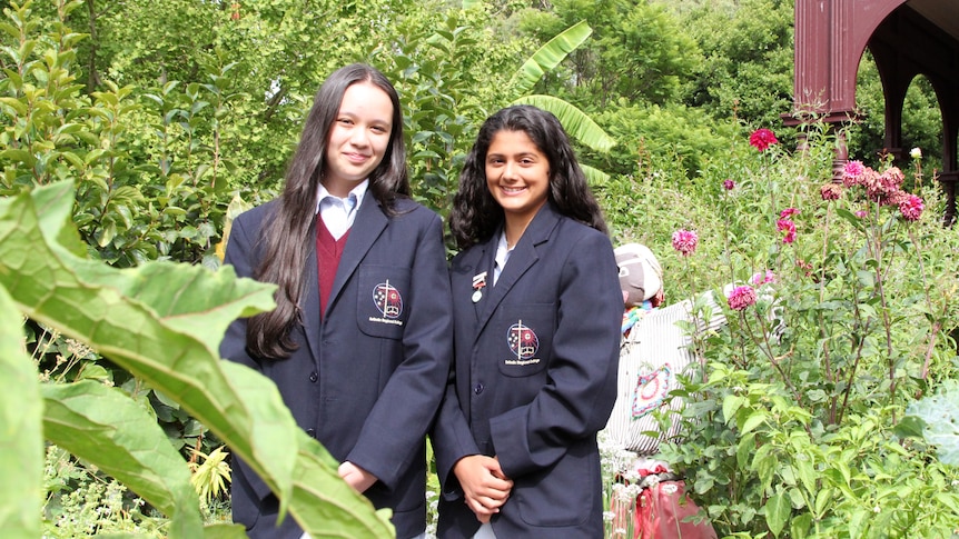 Two schoolgirls with navy blazers standing in a green veggie garden with a maroon building behind them.