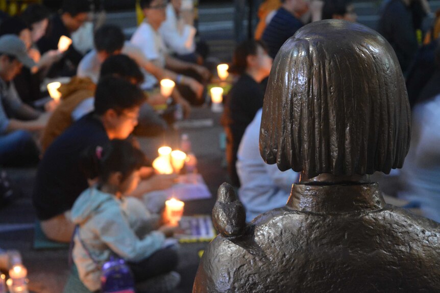 A statues from the back of a woman with short hair and a bird on her shoulder - people with candles sit blurry in the background