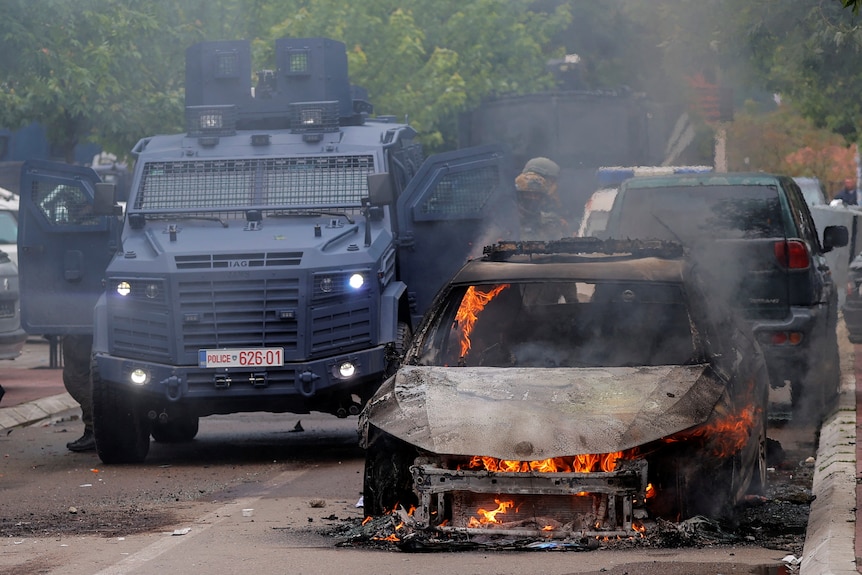 Special police forces officers stand next to a burning car and an armoured vehicle.