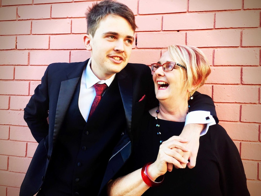 Tall teenager in a suit stands with his arm lovingly around his mother's shoulders while she looks up at him.