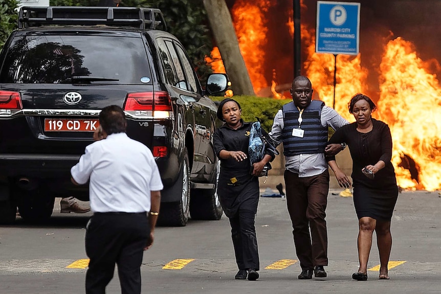 Security forces help civilians flee the scene as cars burn behind.