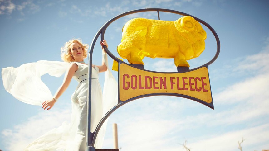 A woman wearing a flowing white dress hangs off a bright yellow Golden Fleece sign.