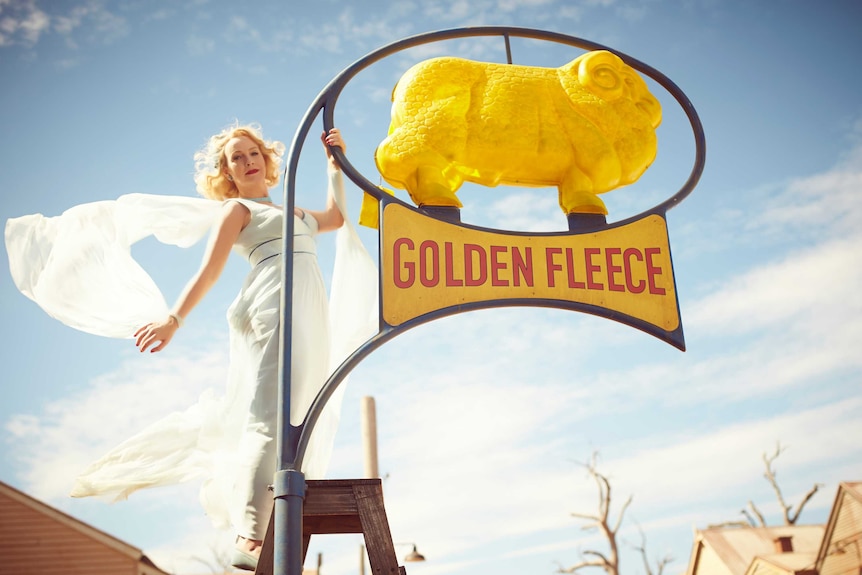A woman wearing a flowing white dress hangs off a bright yellow Golden Fleece sign.