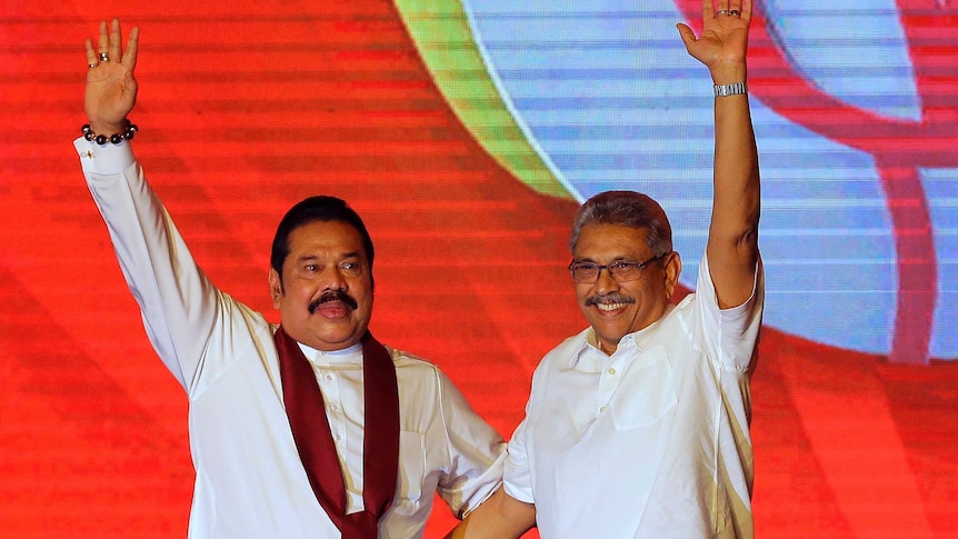 Mahinda Rajapaksa and brother Gotabaya Rajapaksa holds their hands in the air in front of a colourful orange wall.