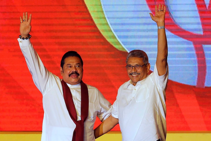 Mahinda Rajapaksa and brother Gotabaya Rajapaksa holds their hands in the air in front of a colourful orange wall.