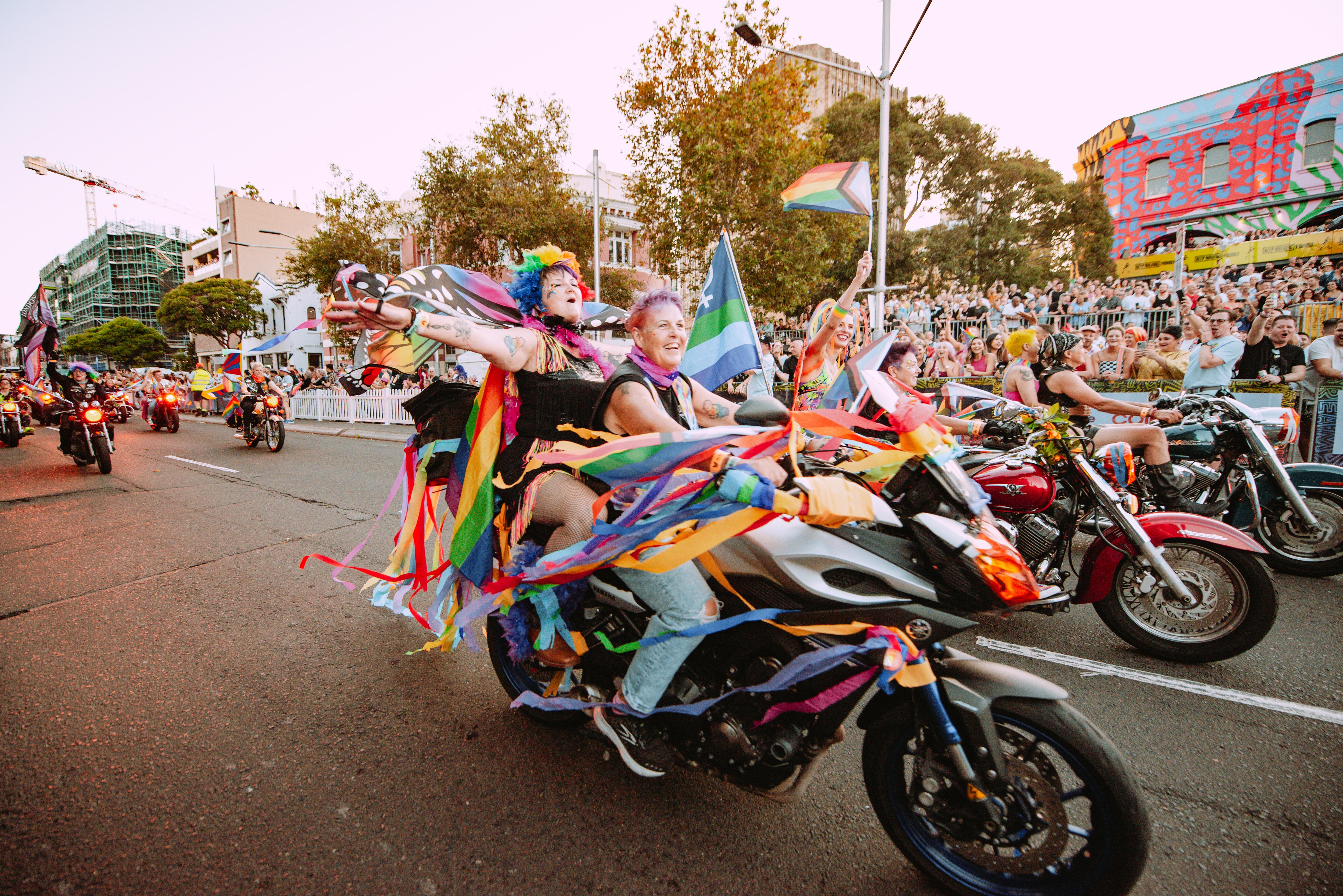 Rainbow flags in the wind attached to motorbikes, with smiling people sitting on them moving down a busy street.