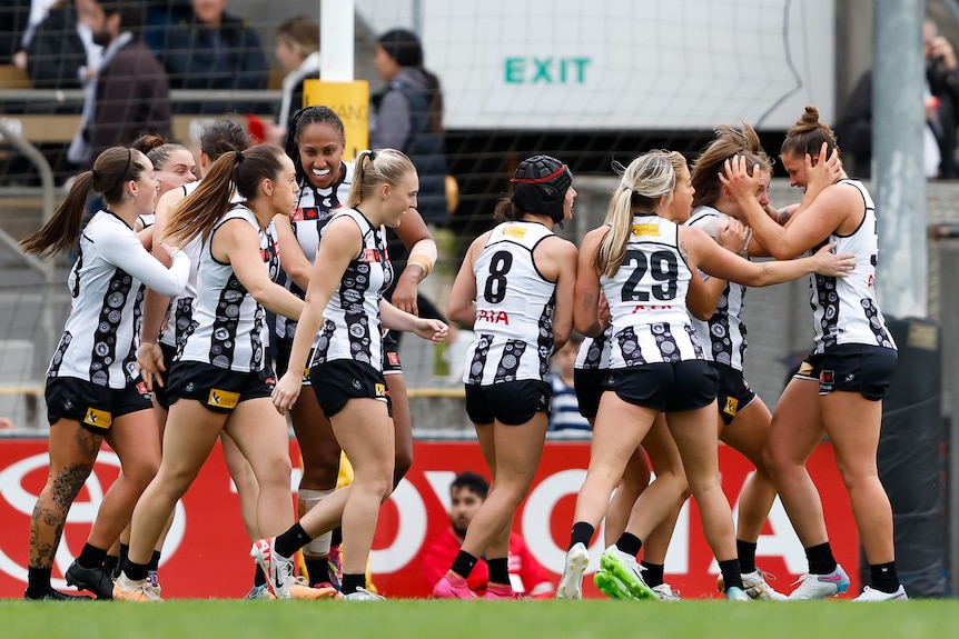A group of Collingwood AFLW players smile and gather to congratulate a goalscorer.