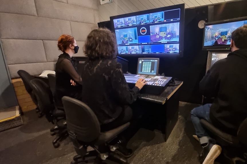 Three masked people sit with their back to the camera, looking at a screen with multiple live shots on it.