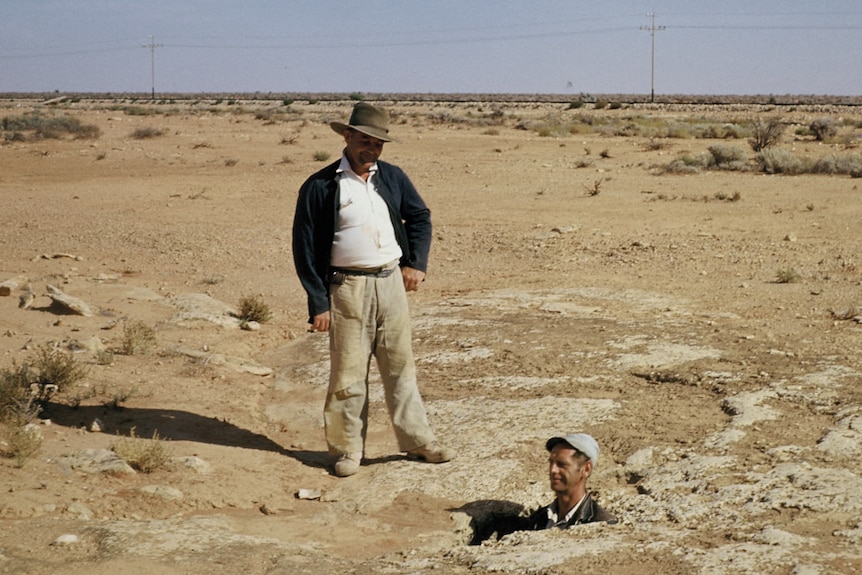 A man stands in a blowhole with just his head exposed with another man looks on