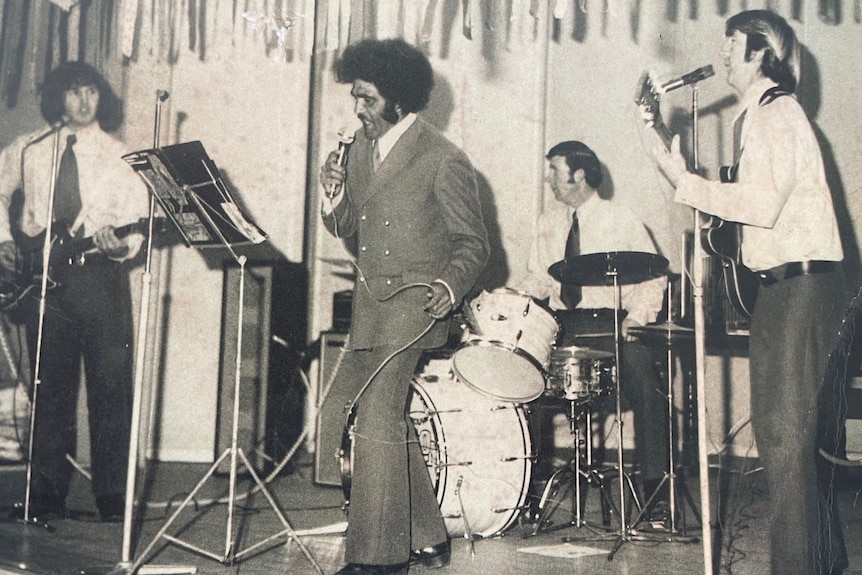 Uncle Herb in the late 60s singing on stage with an afro hairstyle, wears a suit, three Caucasian-looking band members.