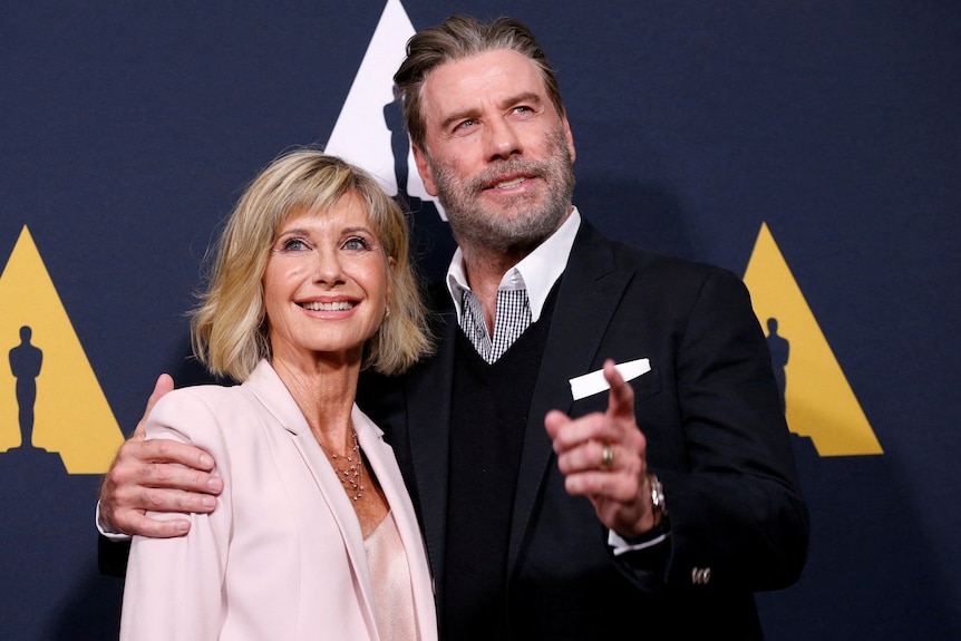 A man and woman smile at the camera. He has his right arm protectively around her shoulders