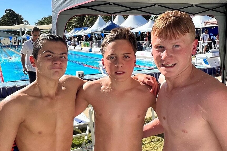 Three teenaged boys with wet hair standing together in front of a pool where a swimming carnival is taking place.