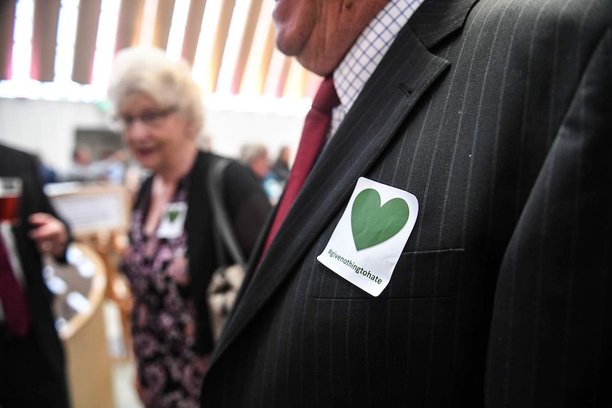 A close-up shot of an elderly man wearing a black and grey pinstriped blazer with a "give nothing to hate" sticker on it.