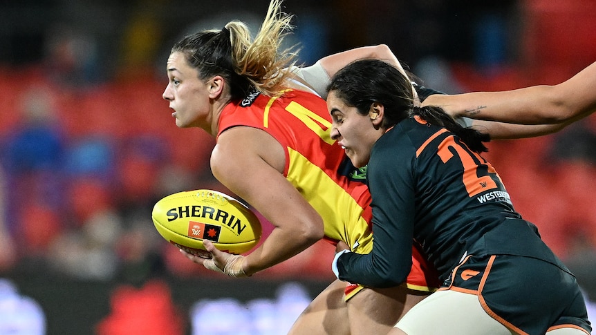 A woman wearing a read and yellow shirt holding a sherrin AFL football and running while another woman wearing black tackles her