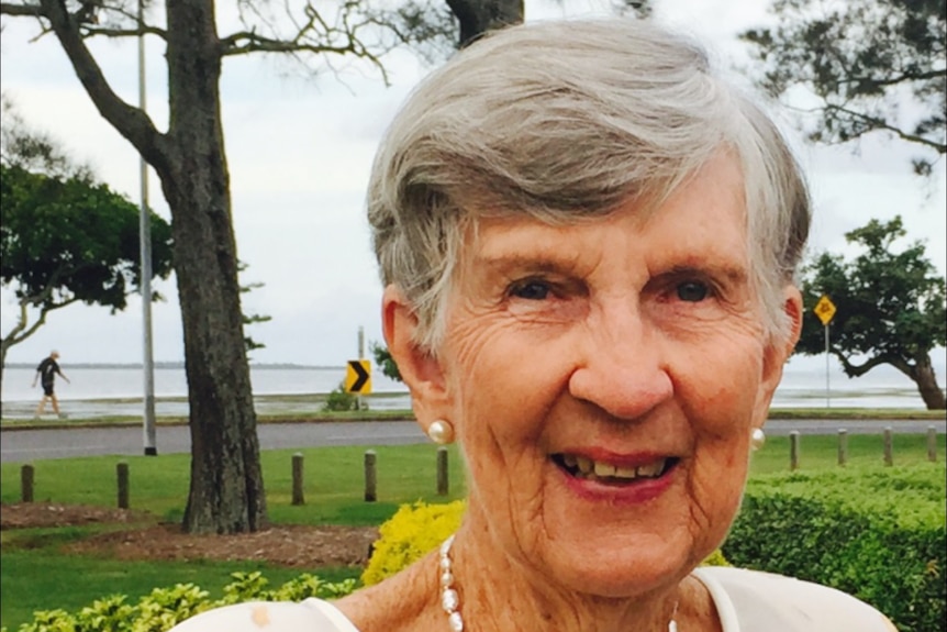 An elderly woman standing in a park in front of the ocean, smiling.