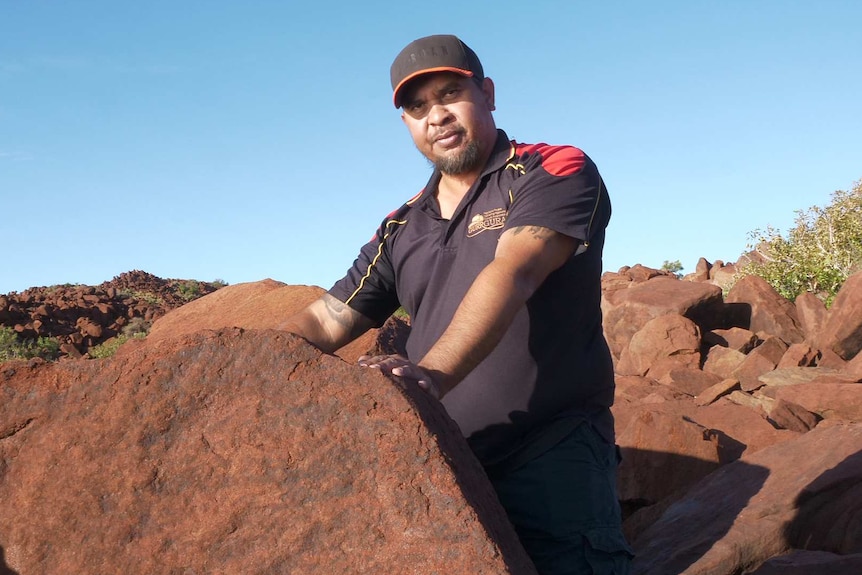 An Indigenous man with a goatee, wearing  a cap and t-shirt, places his hands on red rock.