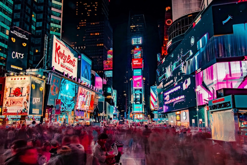 Crowds rush through neon lights and bright signs in Times Square in New York at night.