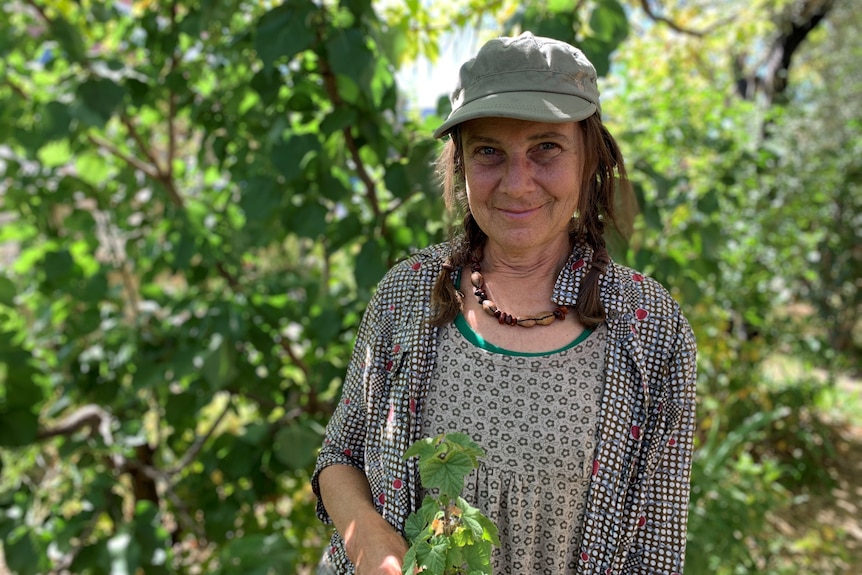 A woman with dark hair and wearing a green cap standing in front of lush green trees at a community garden.