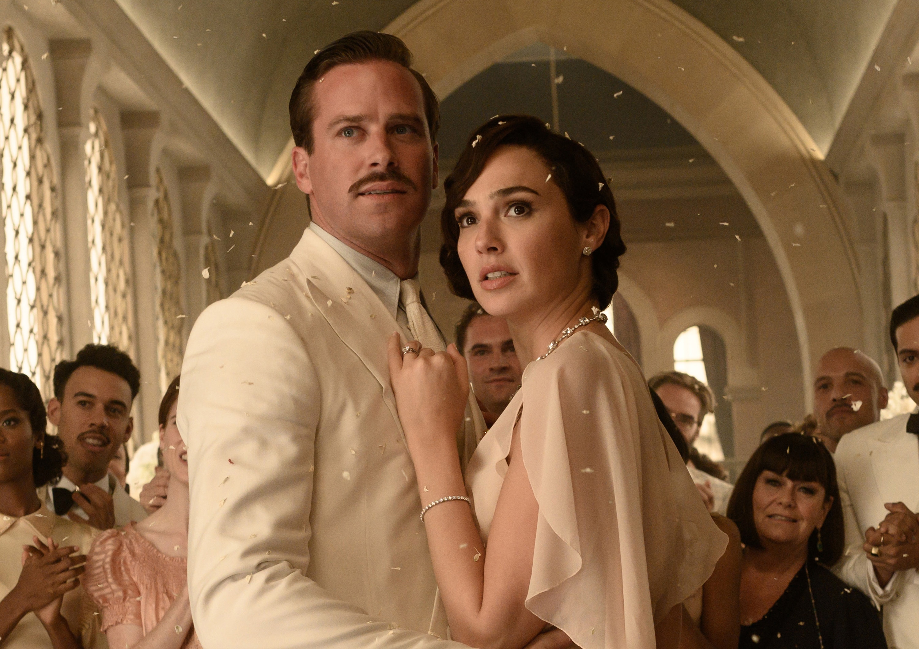 A group of people in black tie dress clap as they surround a troubled looking bride and groom in an ornate space